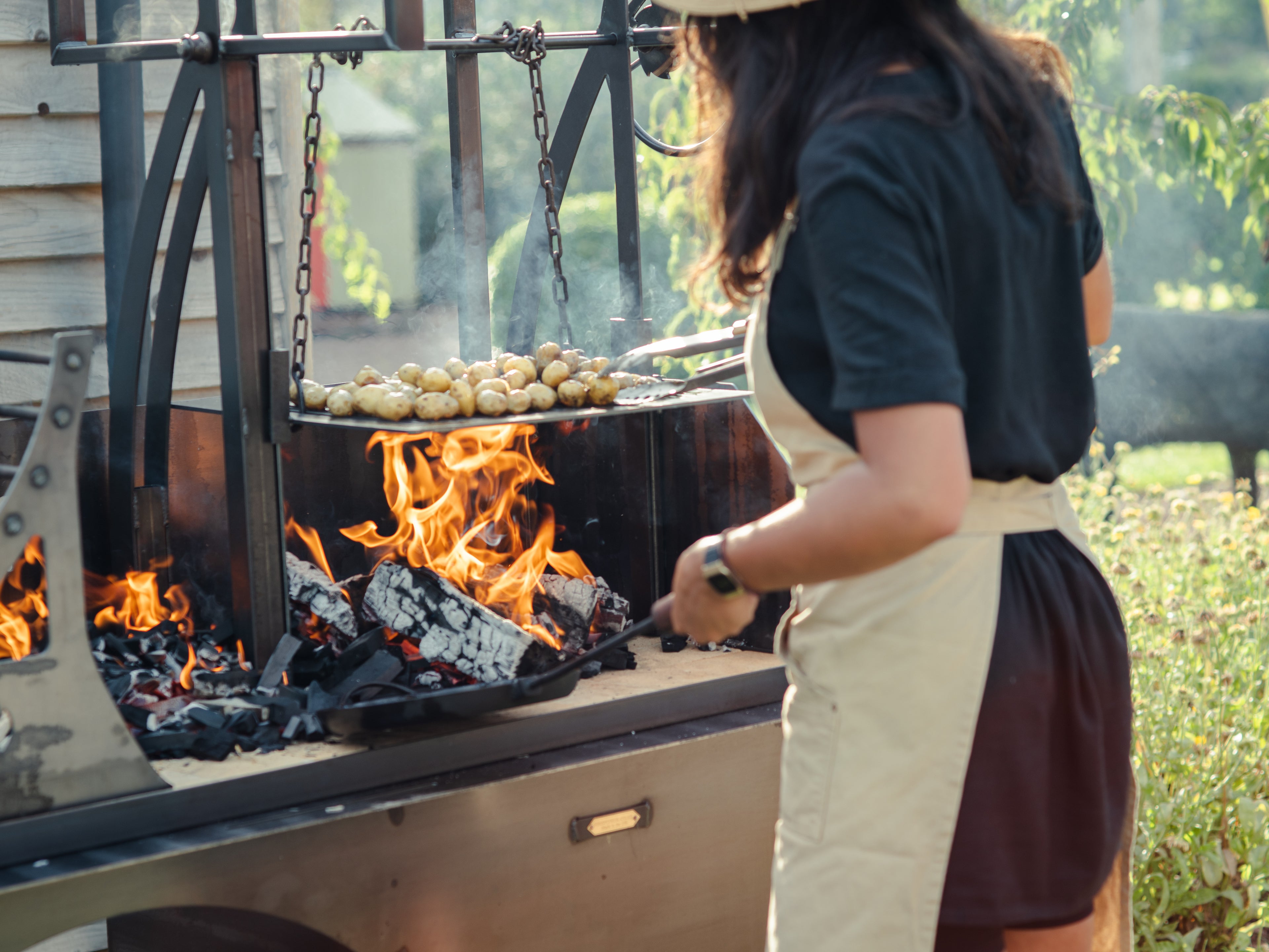 Person cooking potatoes on an outdoor grill with flames, using an Asado fire cage.