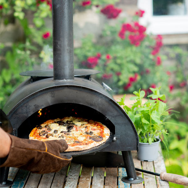 Wood-fired pizza ovens cooking a fresh homemade pizza outdoors in a garden setting with chimney and rustic metal design.